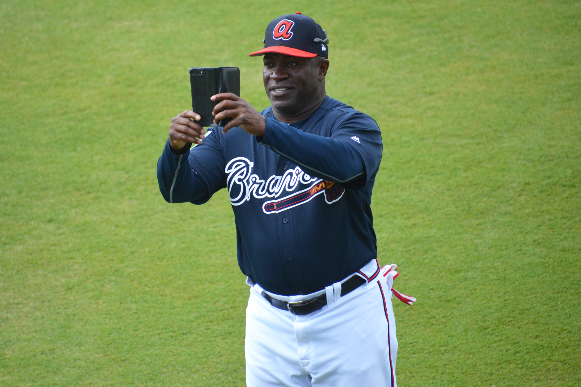 Braves first base coach Eric Young Sr. takes photos of CoolToday Park on his phone during warmups.