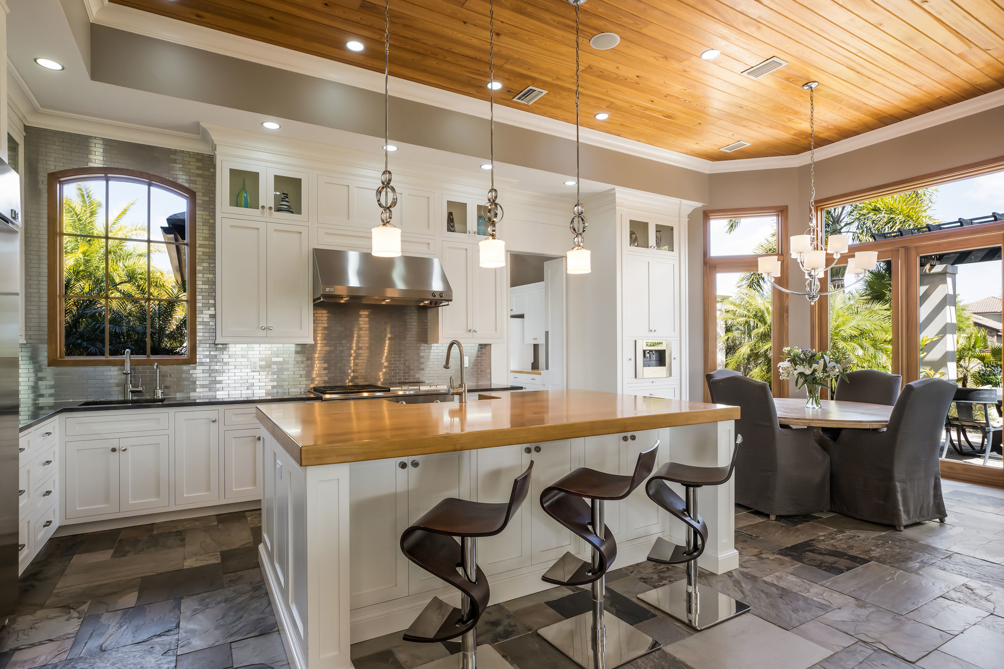 Wood warms up the kitchen with a paneled ceiling and a large island. A breakfast area is to the right, with an outdoor dining terrace through glass doors.