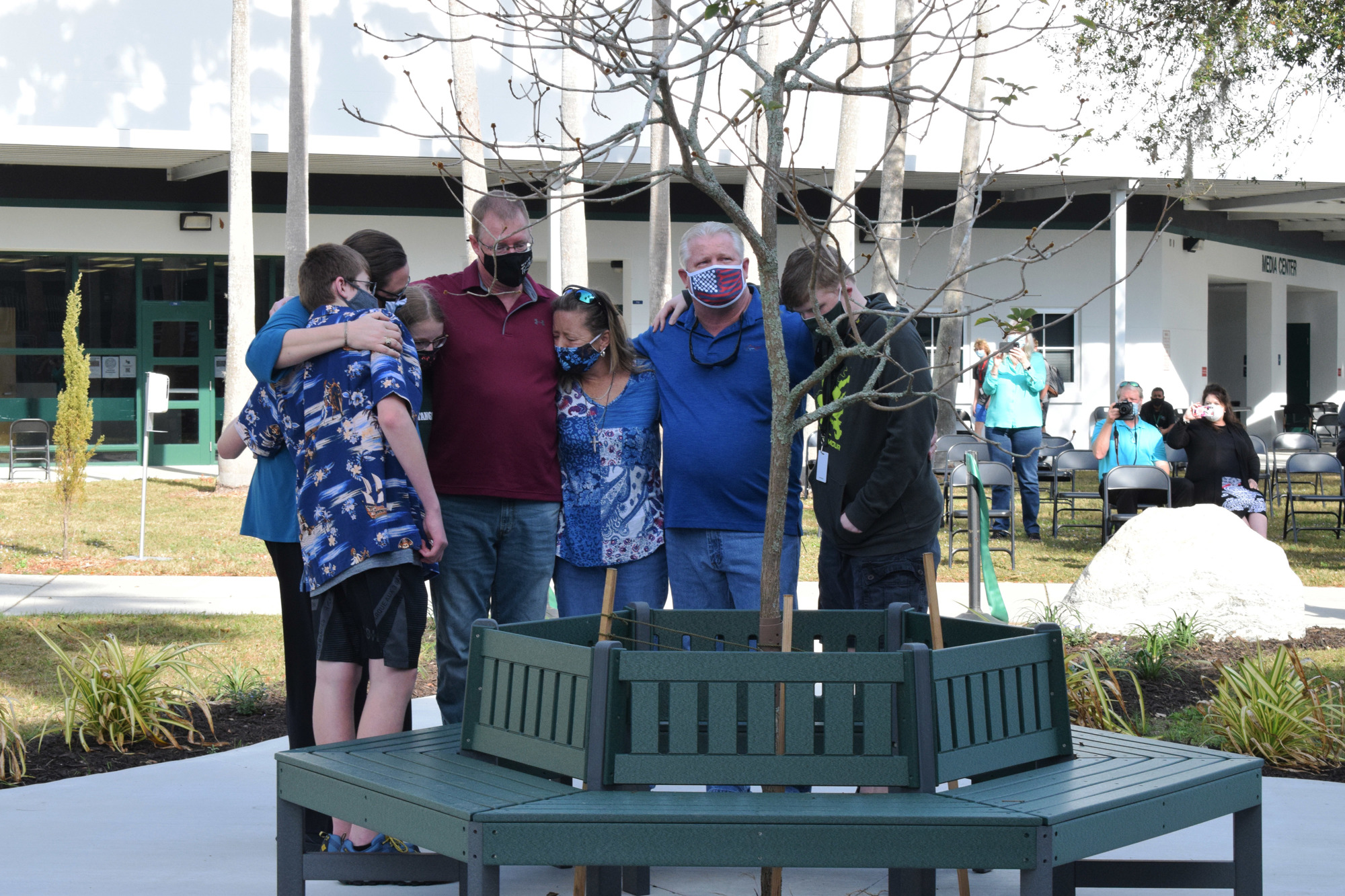 Robert Powers, Rebecca Powers, Katie Powers, Dan Powers, Shayleen Coyner, Scott Coyner and Cory Coyner hug each other while standing in front of the bench that honors their loved ones, Matthew Powers and Chase Coyner.