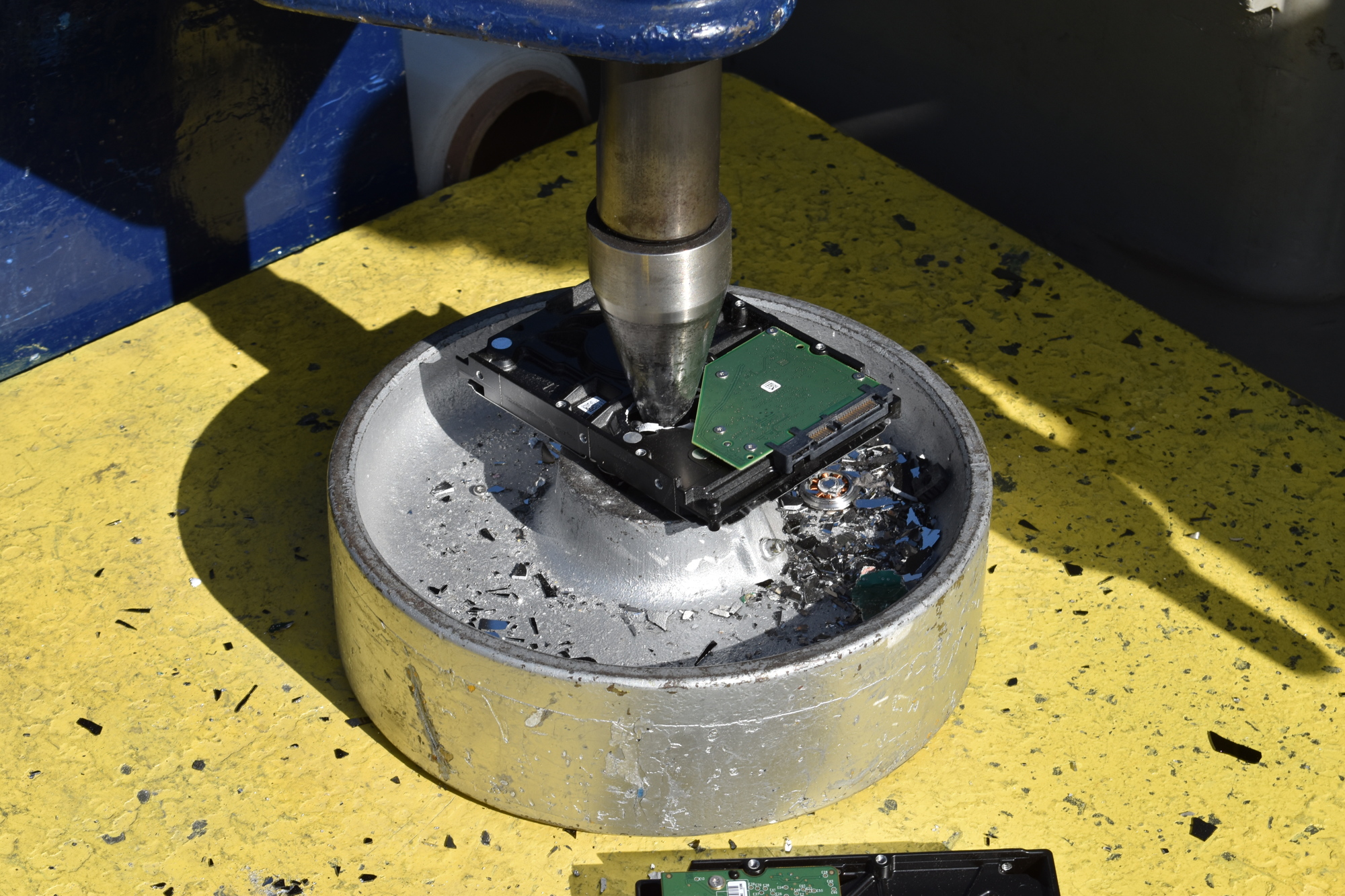 The drill press punctures a hard drive while fragments of hard drive platters litter the surrounding surface.